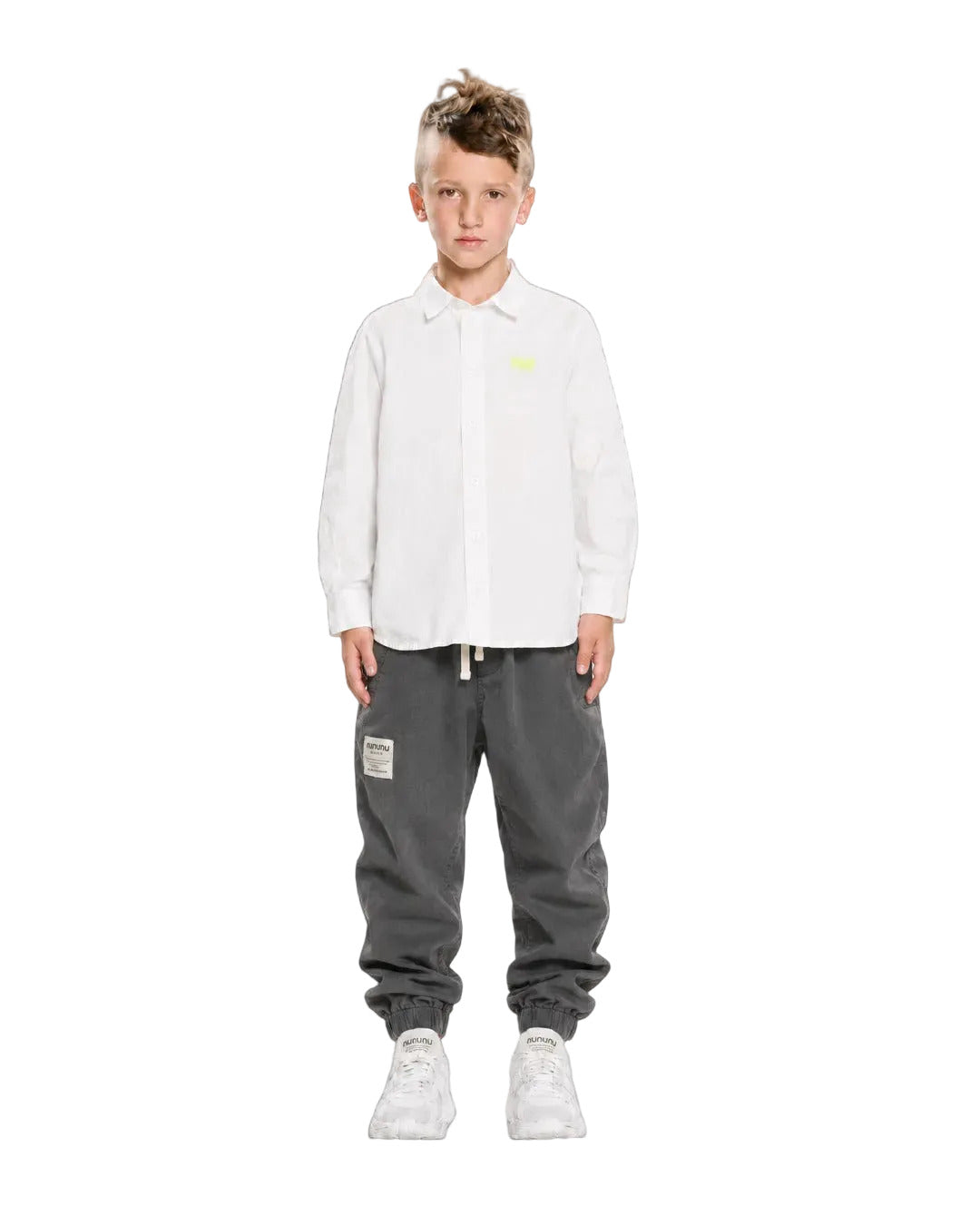 A young boy stands against a plain white studio background wearing a white long-sleeve button-up shirt with a small yellow chest logo, dark gray jogger-style pants with drawstrings and a patch, and white sneakers.