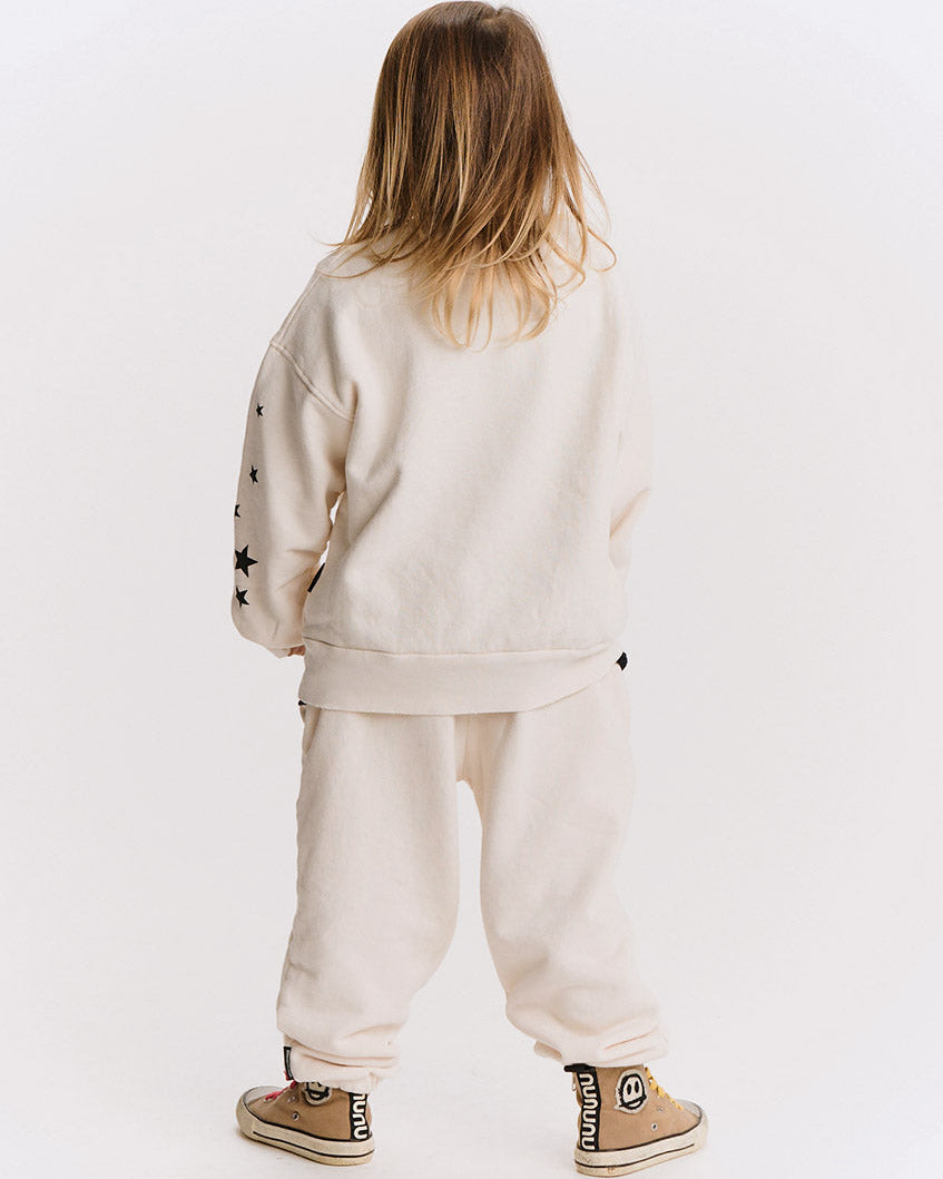 A child viewed from behind wearing a cream-colored sweatshirt and jogger pants set, with black embroidered stars on the sleeve and tan sneakers.