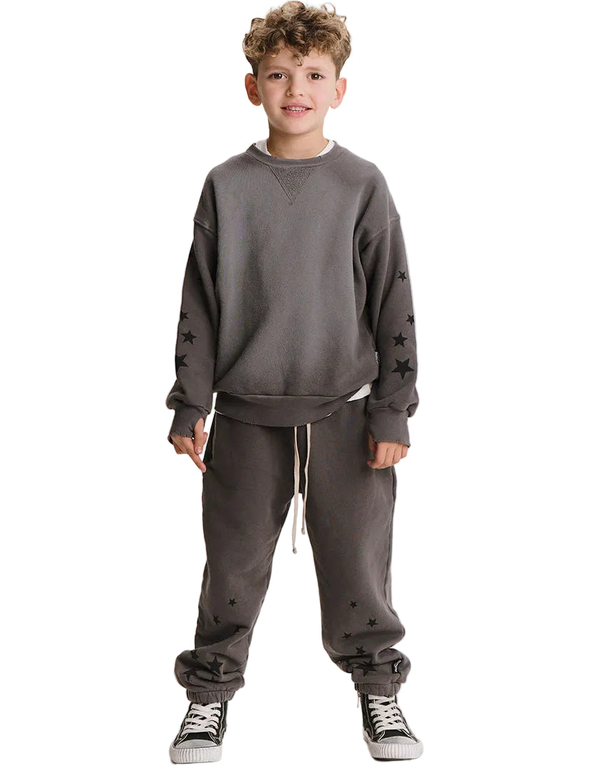 A young boy modeling a dark gray crewneck sweatshirt and matching jogger pants with black star details, paired with black and white sneakers, against a plain white background.