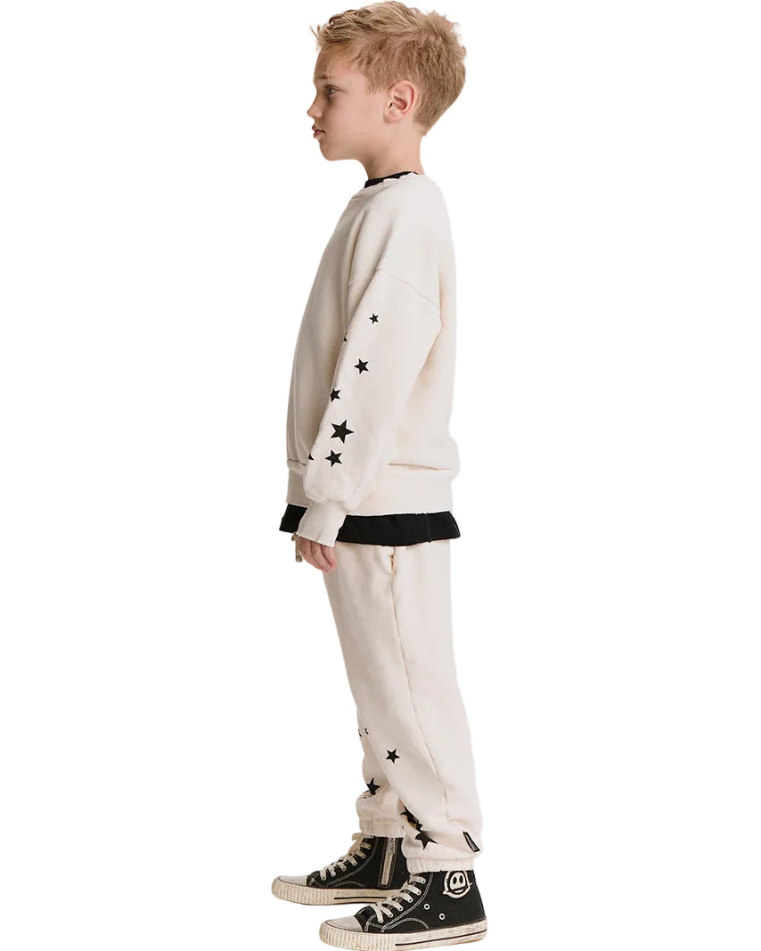A child model shown in profile wearing a cream-colored sweatshirt and matching sweatpants with black star details, layered over a black shirt, and paired with black high-top sneakers against a plain white background.