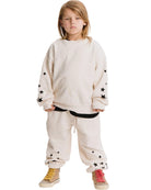 A child model wearing a cream-colored oversized sweatshirt and matching sweatpants, both decorated with black star patterns, paired with yellow and red sneakers, set against a plain white background.