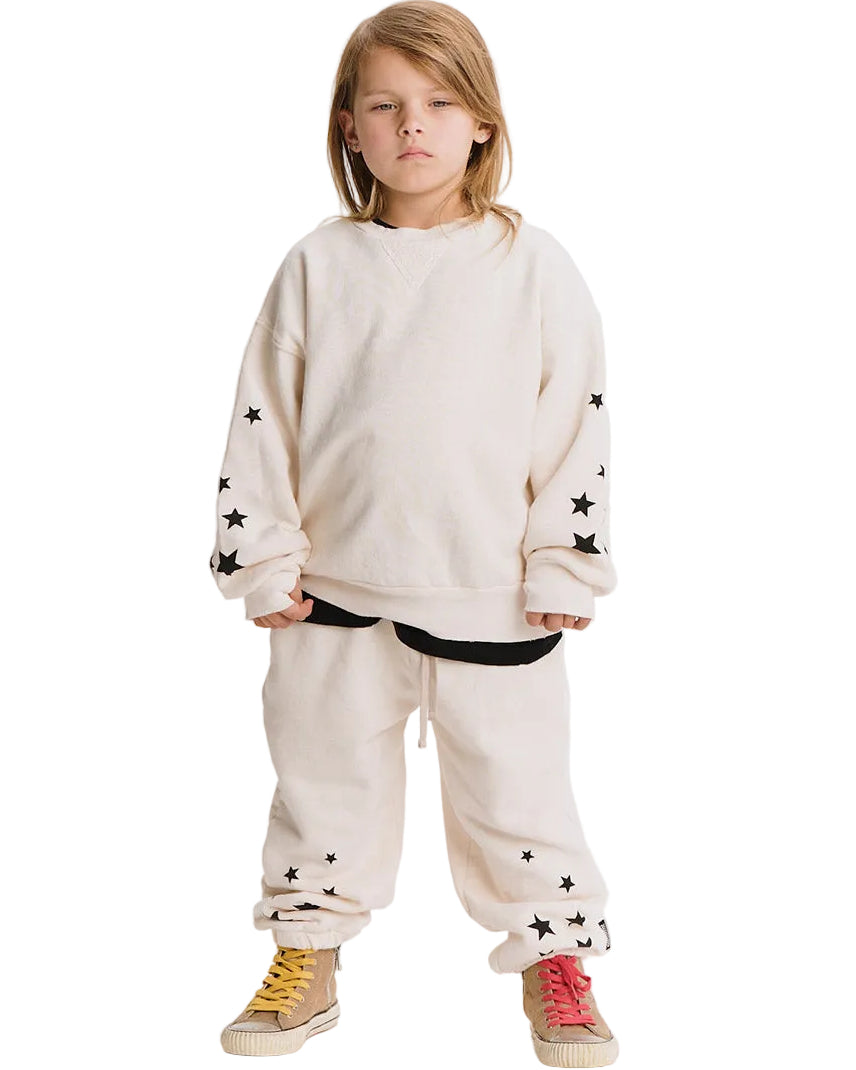 A child model wearing a cream-colored oversized sweatshirt and matching sweatpants, both decorated with black star patterns, paired with yellow and red sneakers, set against a plain white background.