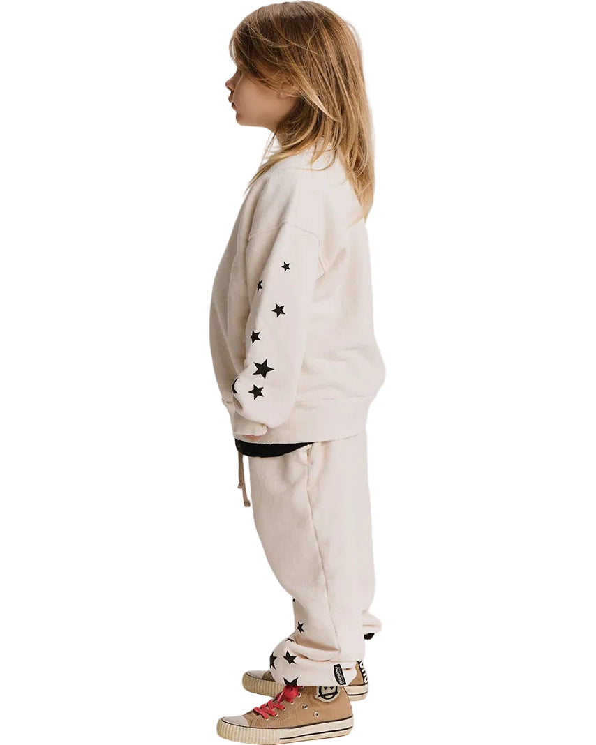 A child model shown in profile wearing a cream-colored two-piece sweatshirt and jogger pants set with black embroidered star details on the sleeves and cuffs, paired with beige sneakers.