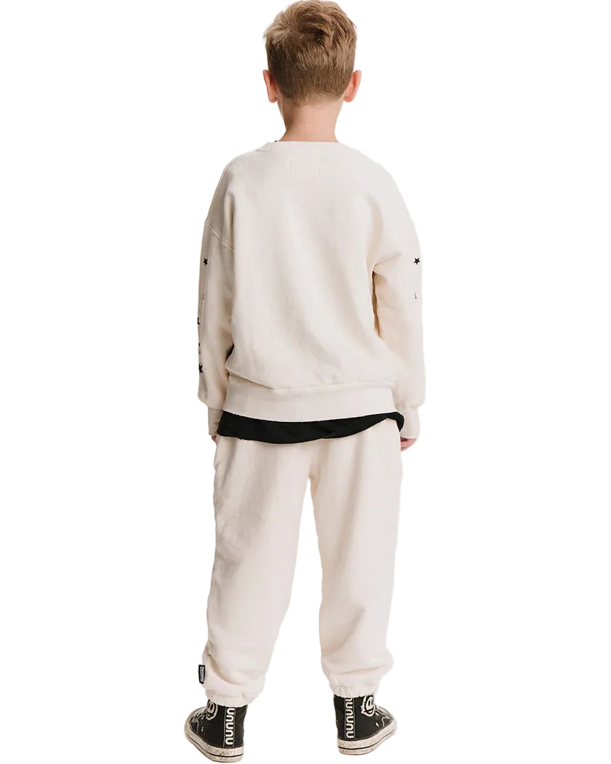 A child viewed from the back wearing a cream-colored sweatshirt and matching sweatpants with a black hem detail, paired with black sneakers.