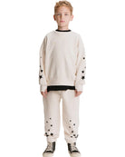 A young boy modeling a cream-colored two-piece sweatshirt and jogger pants set with black star details and black contrast trim, paired with black and white sneakers, on a solid white background.