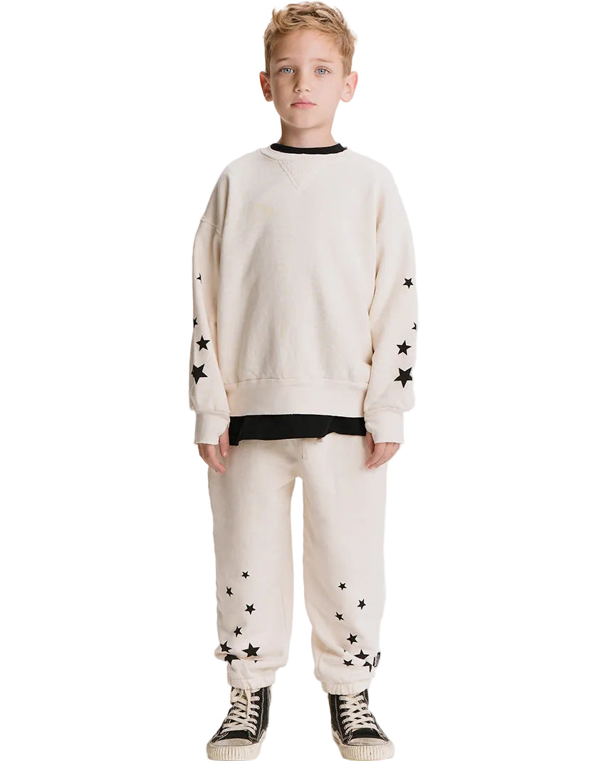 A young boy modeling a cream-colored two-piece sweatshirt and jogger pants set with black star details and black contrast trim, paired with black and white sneakers, on a solid white background.